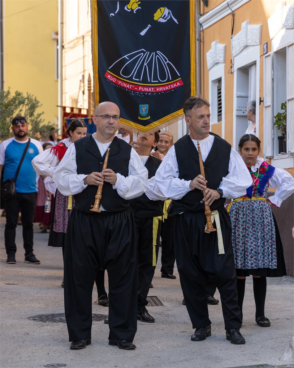 Dva sopca sviraju sopile u povorci ulicama Punta, iza njih barjak KUD-a Punat